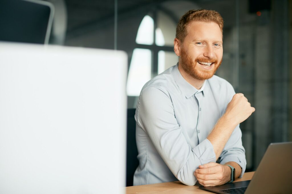 Happy entrepreneur using laptop while working at corporate office and looking at camera.